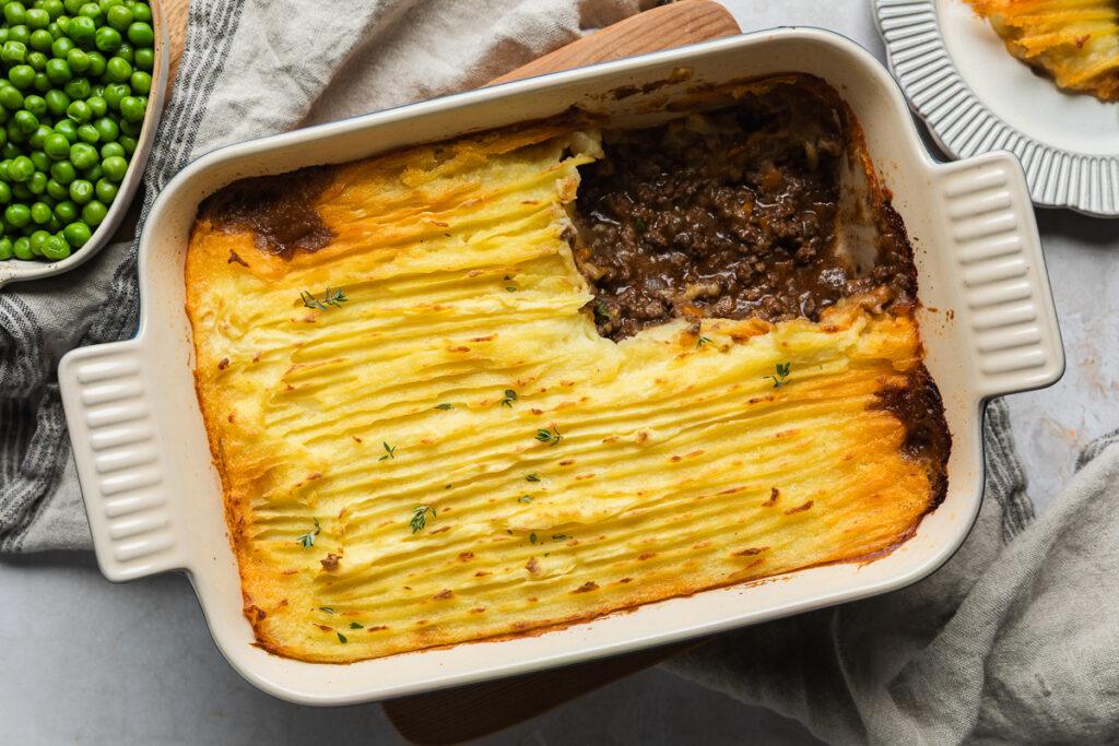 Baked cottage pie in a rectangular dish with a portion of the mashed potato top cut out to show the rich beef filling below.