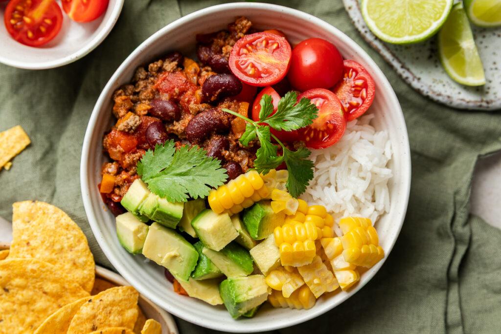 A chilli bowl with rice, topped with avocado, corn and tomatoes and served with corn chips on the side.