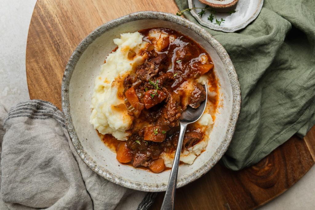 A bowl of slow cooker beef stew and mashed potato with a spoon ready to eat.