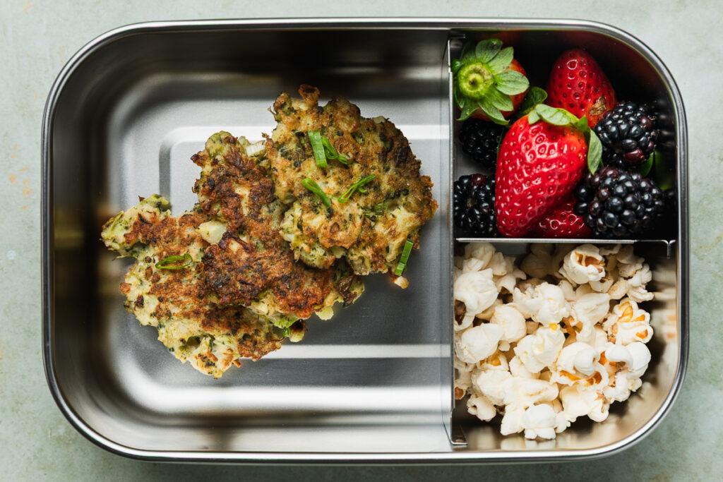 Gluten-free broccoli fritters in a lunchbox with fresh fruit and popcorn.