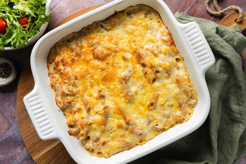 A square baking dish of tuna pasta bake ready to serve, with a bowl of salad just visible in the top left corner.