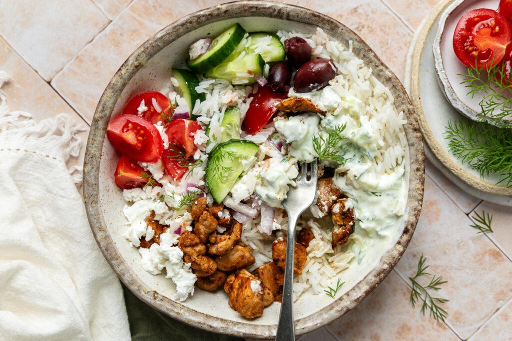 A Greek chicken bowl with the ingredients mixed up and a fork ready to eat.