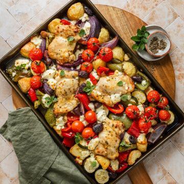A tray of Greek chicken thighs and potatoes on a wooden board.