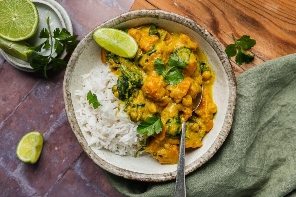 A bowl of slow cooker vegetable curry served with rice and fresh lime, with a spoon ready to eat.