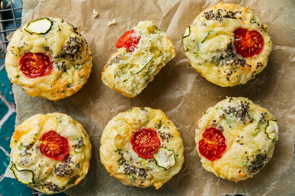 Six zucchini slice muffins on a slice of brown baking paper. Once muffin is halved to show the fluffy texture.