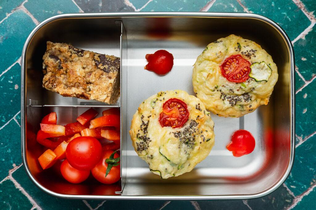 Two gluten-free zucchini slice muffins in a lunchbox with a sweet slice and fresh vegetables.